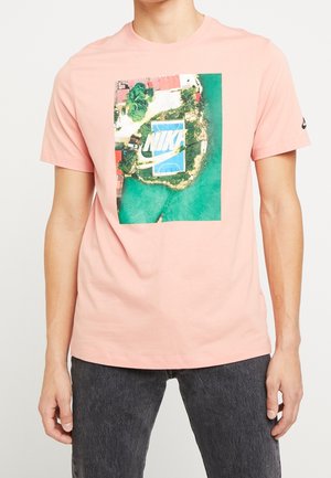 Homme portant un t-shirt rose avec une photo aérienne d'un terrain de basket côtier entouré d'eau et de verdure avec un grand texte blanc "NIKE".