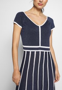 Abito lavorato a maglia blu navy con maniche corte, scollo a V e righe bianche. Presenta un tessuto texturizzato e un corpino aderente che sfocia in una gonna a campana.