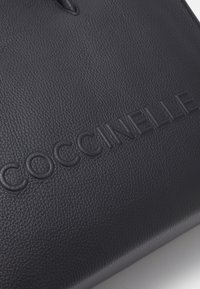 Sac en cuir noir avec une surface texturée et le nom de marque « COCCINELLE » embossé dans le coin inférieur droit. Design minimaliste sans accents supplémentaires.