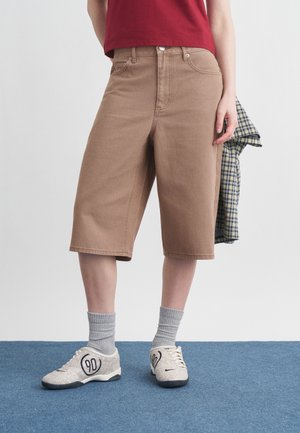 Person iført brune knælange shorts, grå sokker og lyse sneakers med sorte mønstre, der holder en ternet skjorte i den ene hånd.