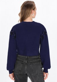 Maglione croppato blu navy con maniche oversize, caratterizzato da dettagli neri con lacci sulle spalle e un orlo a coste. Tessuto a maglia morbida.