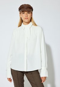 Blusa bianca con dettaglio al collo annodato, maniche lunghe con accenti di bottoni, abbinata a pantaloni marroni aderenti e un cappello di pelle marrone.