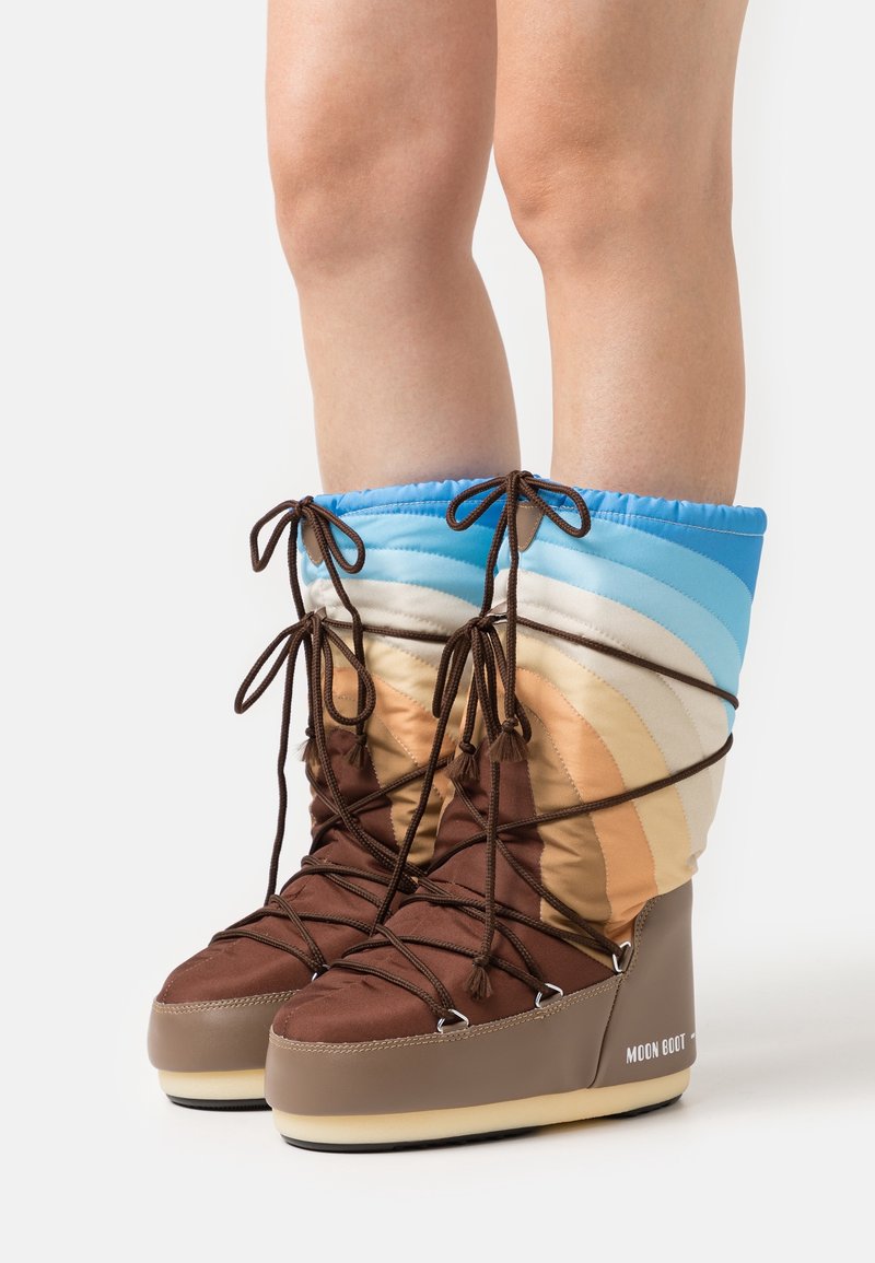 Personne portant des bottes d'hiver rembourrées avec des bouts marron, des tiges dégradées multicolores et des lacets marron enroulés autour des jambes.