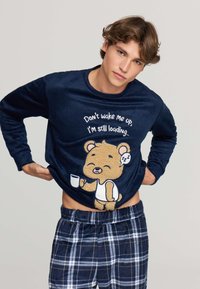 Felpa in pile blu navy con una grafica di un orso e testo. Abbinata a pantaloni da pigiama a quadri blu e bianchi. Texture morbida in tutto.