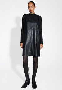 Zwarte leren shift-jurk met een V-hals en twee voorzakken, gedragen over een zwarte geribbelde top, gecombineerd met zwarte enkelboots.