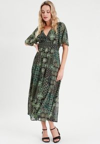 Robe maxi en tissu transparent à motif floral vert, avec un décolleté en V, des manches courtes flottantes et une taille cintrée, associée à des talons noirs.