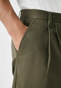 Olive green trousers made of smooth fabric with a pleated design, featuring a side pocket and a flat waistband.