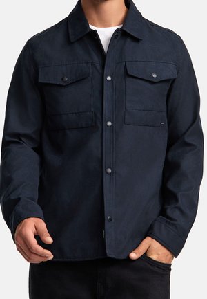 Mann trägt eine marineblaue Jacke mit Knopfleiste, zwei Brustpatten und darunter ein weißes Hemd.