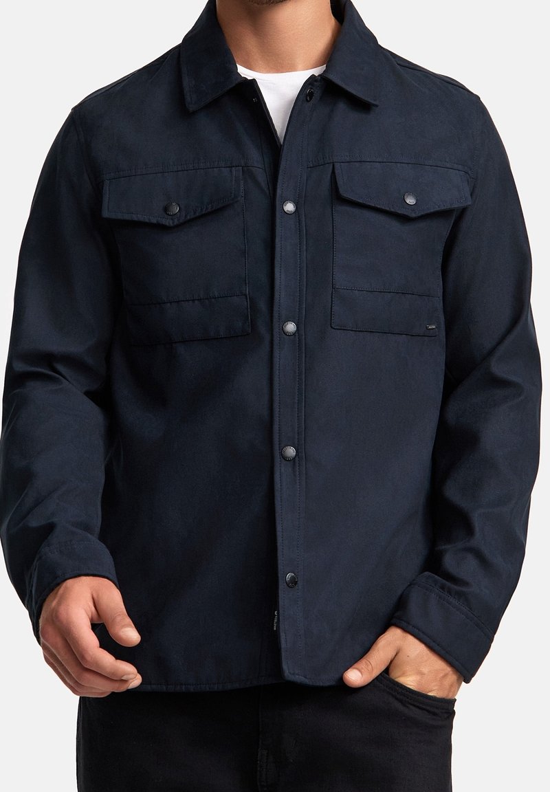 Homme portant une veste bleu marine boutonnée avec deux poches à rabat sur la poitrine et une chemise blanche en dessous.