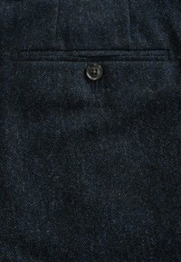 Ciemnoniebieska tkanina o fakturze mieszanki wełny z subtelnym wzorem. Zawiera pojedynczy czarny guzik oraz detal tylnej kieszeni.