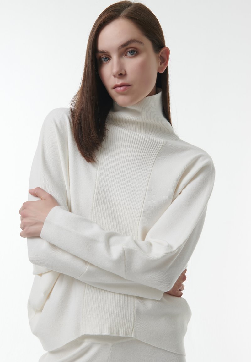 Witte coltrui met geribbelde textuur en een relaxte pasvorm. Beschikt over brede mouwen en een subtiele naaddetail langs de voorkant.