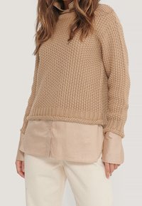 Beige gestrickter Pullover mit lockerer Passform und strukturiertem Muster, darüber ein hellbeige Hemd mit Knöpfen. Hellcremefarbene Hose rundet das Outfit ab.