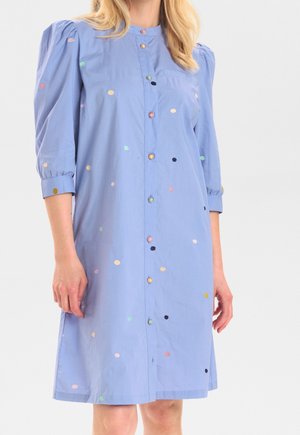 Mujer con vestido azul claro hasta la rodilla con mangas abullonadas tres cuartos y lunares multicolores, de pie sobre un fondo liso.