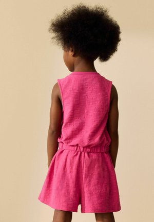 Child standing facing away wearing textured sleeveless pink top and matching shorts against beige background.