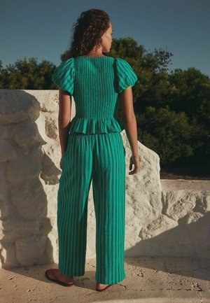 Femeie cu păr creț purtând un top cu dungi în nuanță teal cu mâneci bufante și pantaloni cu picior larg, stând în aer liber lângă un zid de piatră cu copaci pe fundal.