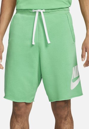 Mężczyzna ubrany w jasnooliwkowe spodenki Nike z białym sznurkiem i bocznymi kieszeniami, stojący z rękami swobodnie opuszczonymi wzdłuż ciała.