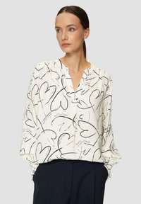 Blouse blanche avec des motifs de cœurs noirs et un texte écrit, présentant un col en V, des manches longues et des poignets froncés. Tissu léger et fluide.