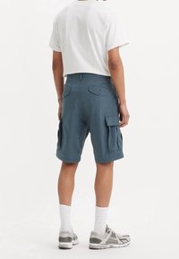 Muži v modrých cargo šortkách, bílých ponožkách, šedých a bílých sportovních botách a bílém tričku, stojící zády k divákovi.