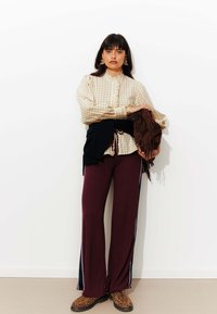Femme se tient contre un mur blanc, portant une blouse à carreaux beige, un pantalon de survêtement bordeaux avec des rayures sur le côté, des bottes à imprimé léopard, tenant un sac à franges marron.