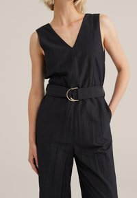 Czarny kombinezon z teksturowanej tkaniny, z głębokim dekoltem w kształcie litery V, bez rękawów, z wcięciem w talii i ozdobą w postaci złotego, okrągłego klamry.