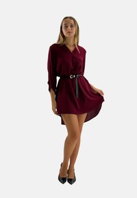 Robe bordeaux avec un décolleté en V et des manches longues, cintrée à la taille par une ceinture noire. Tissu texturé et ourlet asymétrique. Talons noirs.