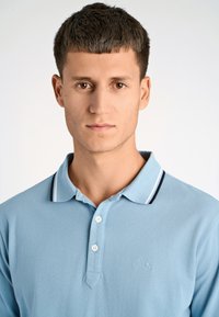 Jasnoniebieska koszulka polo z teksturowanej tkaniny, z dwoma guzikami, kontrastowym kołnierzykiem z ciemnymi paskami oraz subtelnym haftowanym logo.