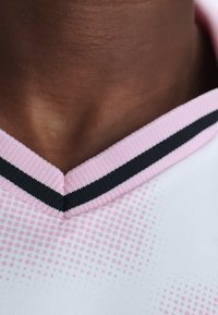 Koszulka z dekoltem w kształcie litery V z różowym i czarnym wykończeniem noszona przez osobę o ciemnym kolorze skóry; koszulka ma różowy wzór w kropki na białym materiale.