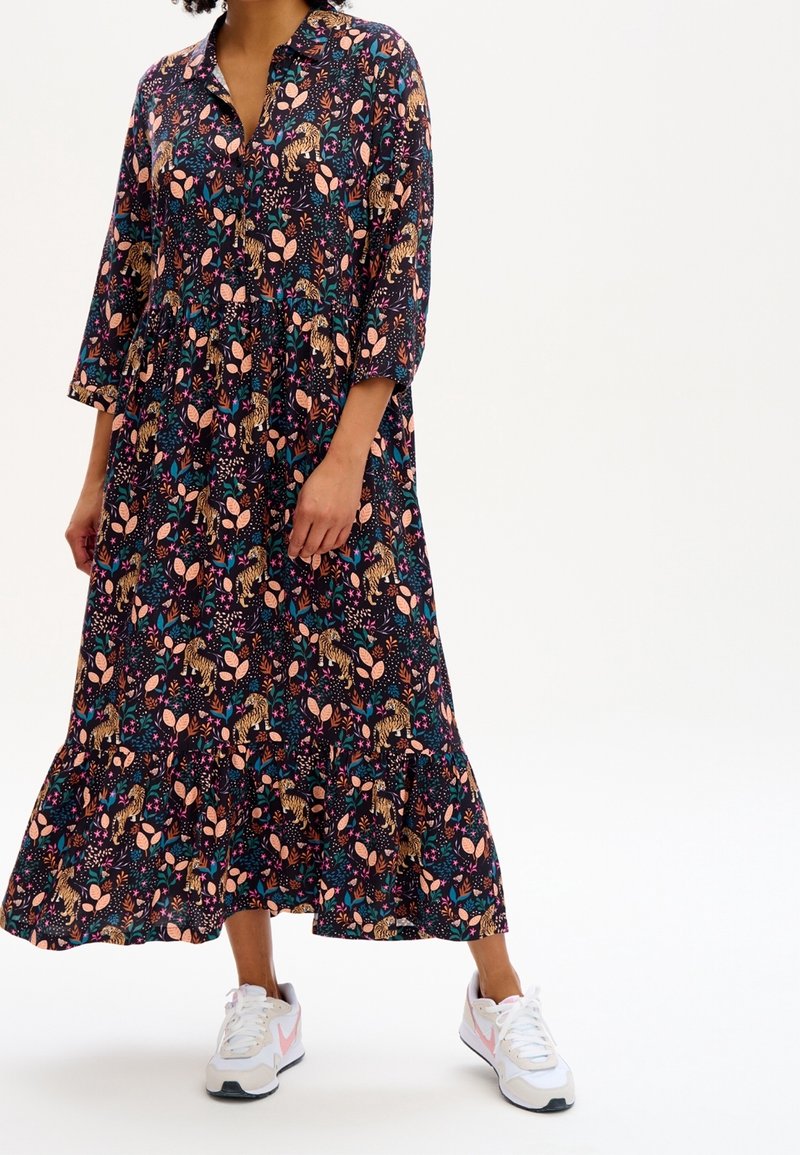 Langes, dunkles Blumenkleid mit Blatt- und Tigermustern, das einen Kragen und dreiviertellange Ärmel hat. Getragen mit weißen Sportschuhen.