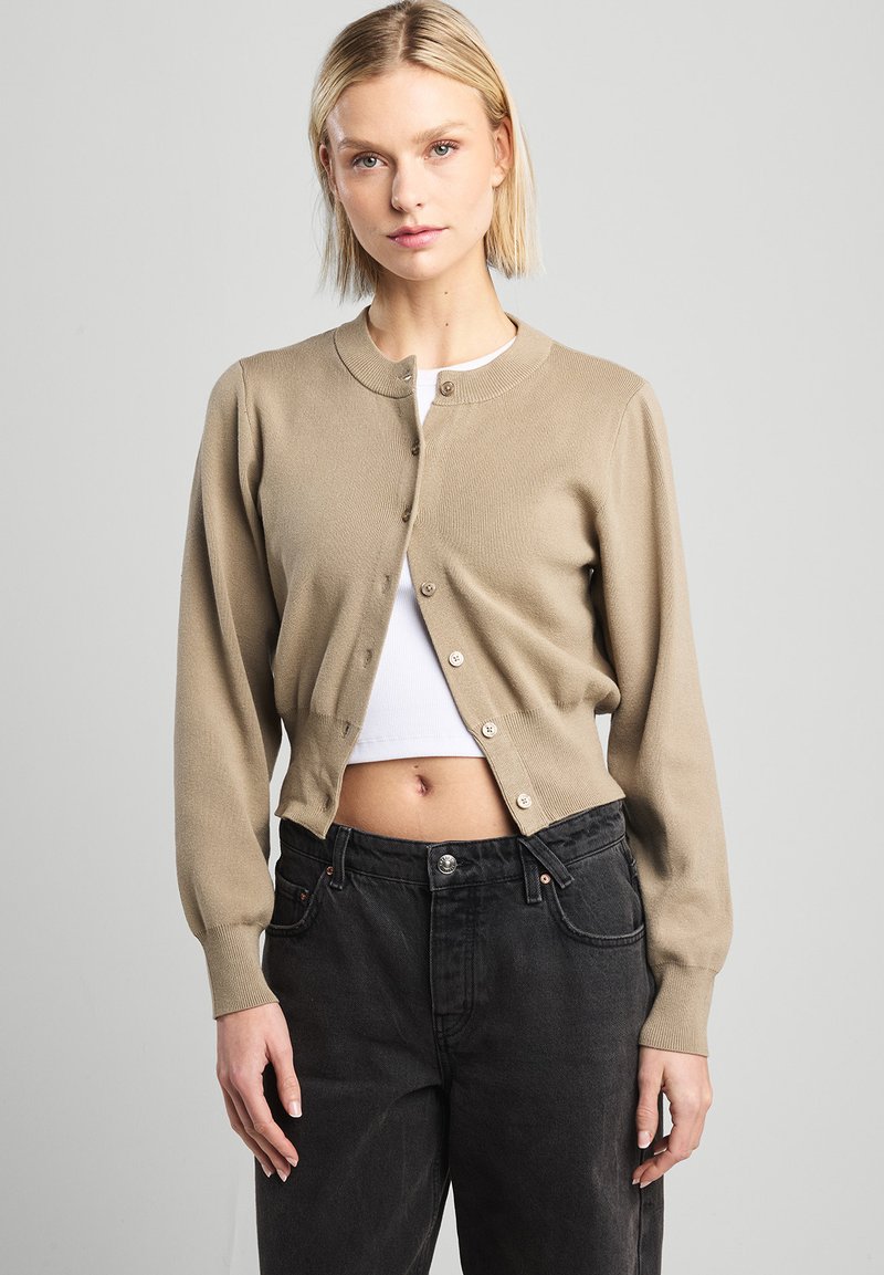 Beige knitted cardigan with a round neckline and long sleeves, featuring five front buttons, paired with high-waisted black jeans.