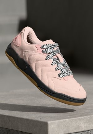 Lyserøde ruskinds sneakers med en tekstureret overdel, sort og hvid mønstrede snørebånd og en gummisål med en farvetudsida i beige.