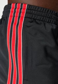 Black athletic shorts featuring a textured fabric, elastic waistband, and three vertical stripes in red, white, and black on the side.