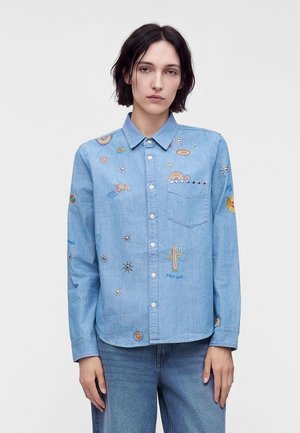 Γυναίκα που φοράει ανοιχτό μπλε denim πουκάμισο με κουμπιά και πολύχρωμα κεντημένα σχέδια, όπως κάκτοι, ουράνια τόξα και αστέρια, συνδυασμένο με μπλε τζιν.
