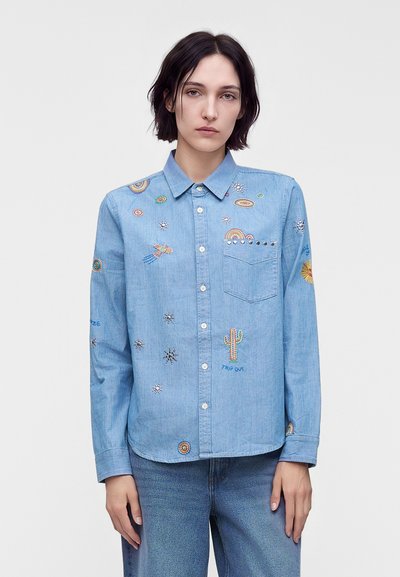 Γυναίκα που φοράει ανοιχτό μπλε denim πουκάμισο με κουμπιά και πολύχρωμα κεντημένα σχέδια, όπως κάκτοι, ουράνια τόξα και αστέρια, συνδυασμένο με μπλε τζιν.