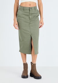 Khaki green knee-length skirt made of cotton with a front slit, featuring belt loops and button closure. Paired with brown ankle boots.