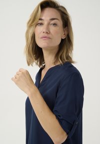 Granatowa bluzka z dekoltem w serek i podwiniętymi rękawami, wykonana z gładkiego materiału, z delikatnym połyskiem i czystym, minimalistycznym designem.