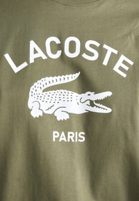 Olivno zeleno blago z belim logotipom Lacoste, ki vključuje grafiko krokodila z odprtimi usti in besedo "Pariz" spodaj.