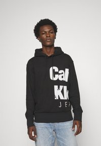 Felpa nera con una grande stampa di testo bianco. Presenta un cappuccio con lacci e polsini a coste. Abbbinata a jeans azzurri chiari.