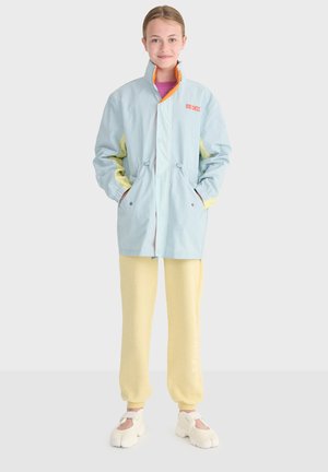 Mladá osoba stojaca, oblečená v svetlomodrej a žltej bunde, žltých nohaviciach a bielych topánkach s rozdelenými špičkami, na jednofarebnom svetlom pozadí.