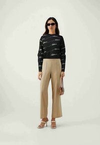 Black sweater with white logo print, beige high-waisted trousers, and heeled sandals. Accessories include small handbag and sunglasses.