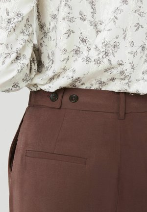 Pantalons marron ajustés avec une texture lisse, dotés de deux boutons noirs à la taille et d'une poche arrière.