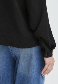 Schwarze Bluse mit langen Ärmeln und strukturiertem Finish, mit geknöpften Manschetten. Kombiniert mit hellblauen weit geschnittenen Jeans.