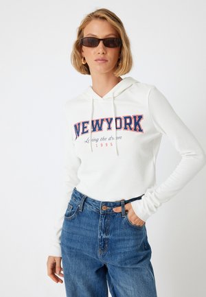 Sweat à capuche blanc côtelé avec le texte "NEW YORK" en rouge et bleu marine, associé à un jean bleu taille haute. Modèle portant des lunettes de soleil noires.