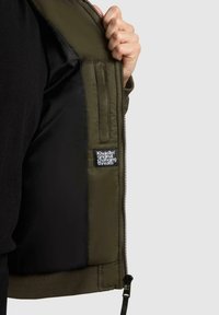 Eine Hand hält eine grüne Jacke offen, die ein schwarzes Innenfutter und ein kleines angenähtes Etikett mit weißem Text zeigt.