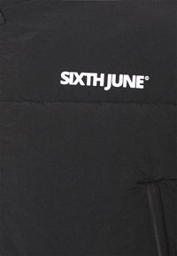 Giubbotto piumino nero con texture liscia, leggero lucido e logo "SIXTH JUNE" bianco sul davanti. Dettaglio della tasca laterale con zip visibile.