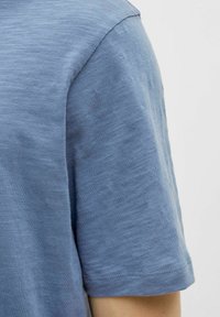 Rövid ujjú ing világoskék anyagból, amely texturált mintával és kerek nyakkivágással rendelkezik, a szélek mentén pedig gondosan varrott részletek találhatók.