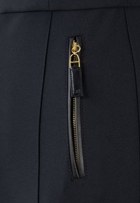 Tessuto nero con una superficie liscia, caratterizzato da una chiusura laterale con zip in metallo dorato e un dettaglio in pelle sul tirazip.