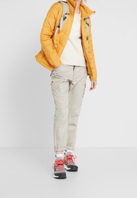 Giacca arancione gonfiata, felpa crema, pantaloni cargo beige chiaro e scarpe da ginnastica nere con accenti rossi, caratterizzati da materiali testurizzati e cuciture visibili.