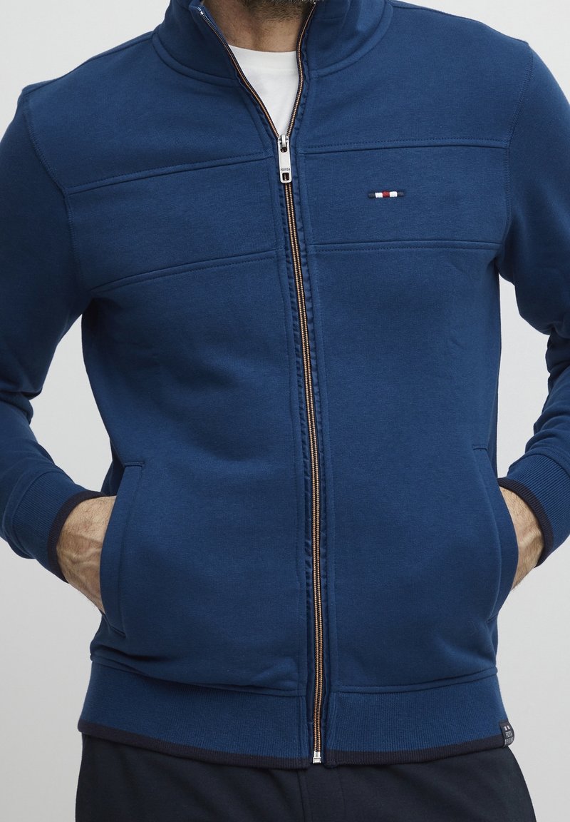 Homme portant une veste bleu marine zippée avec les mains dans les poches avant, t-shirt blanc visible, et petit logo sur la poitrine gauche.