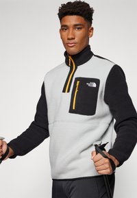 Fleece pullover in grijs en zwart met een ritskraag, een voorzak met een gele rits en een logo-detail op de borst. Geschikt voor het vasthouden van wandelstokken.