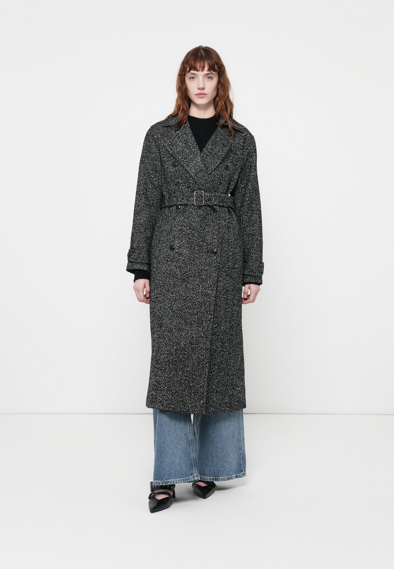Manteau long à motif pied-de-poule gris avec une taille ceinturée, un devant double boutonnage et des revers larges, associé à un jean en denim à jambes larges et des chaussures noires.